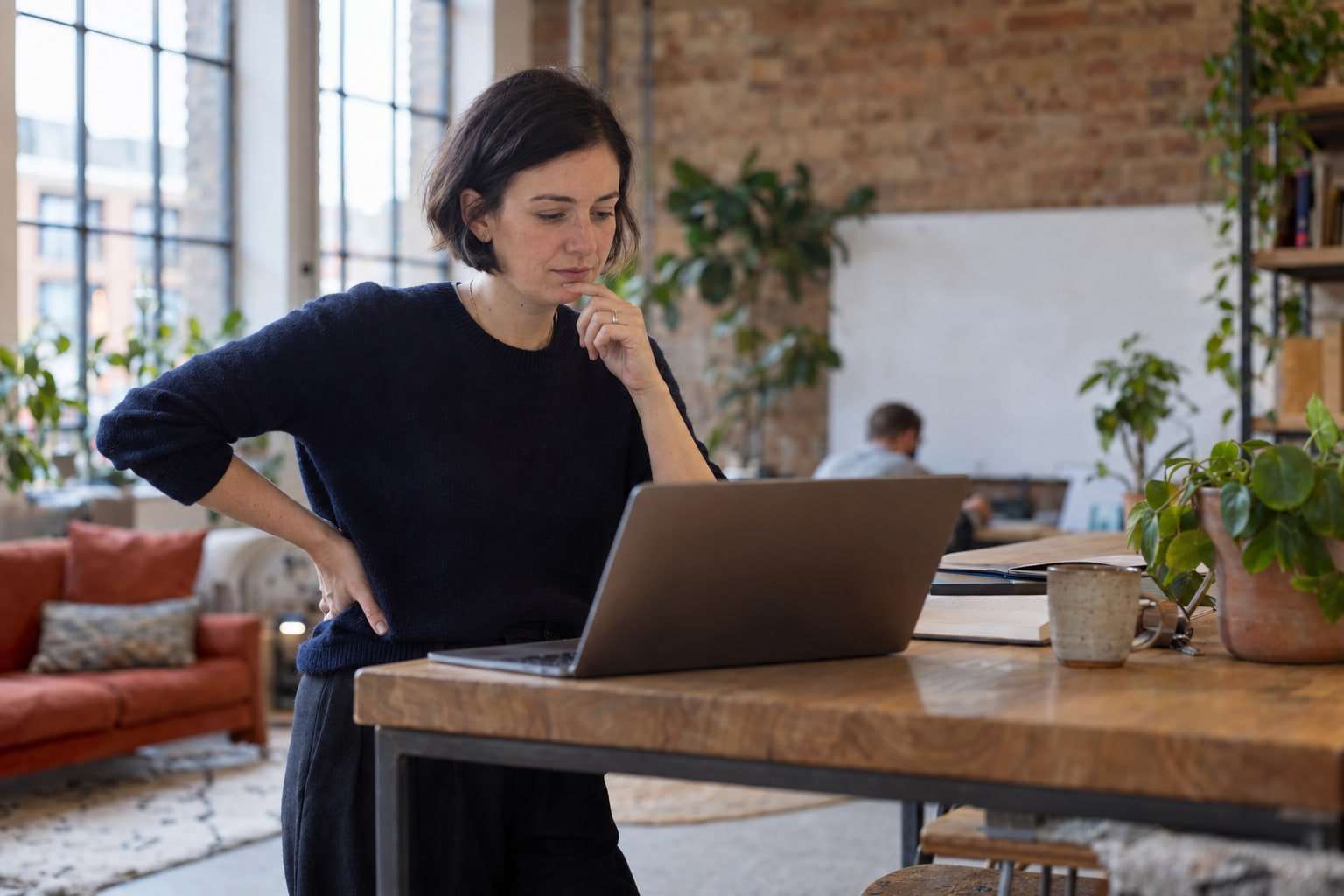 A woman standing at a table in a bright co-working space, thinking as she reads her laptop.