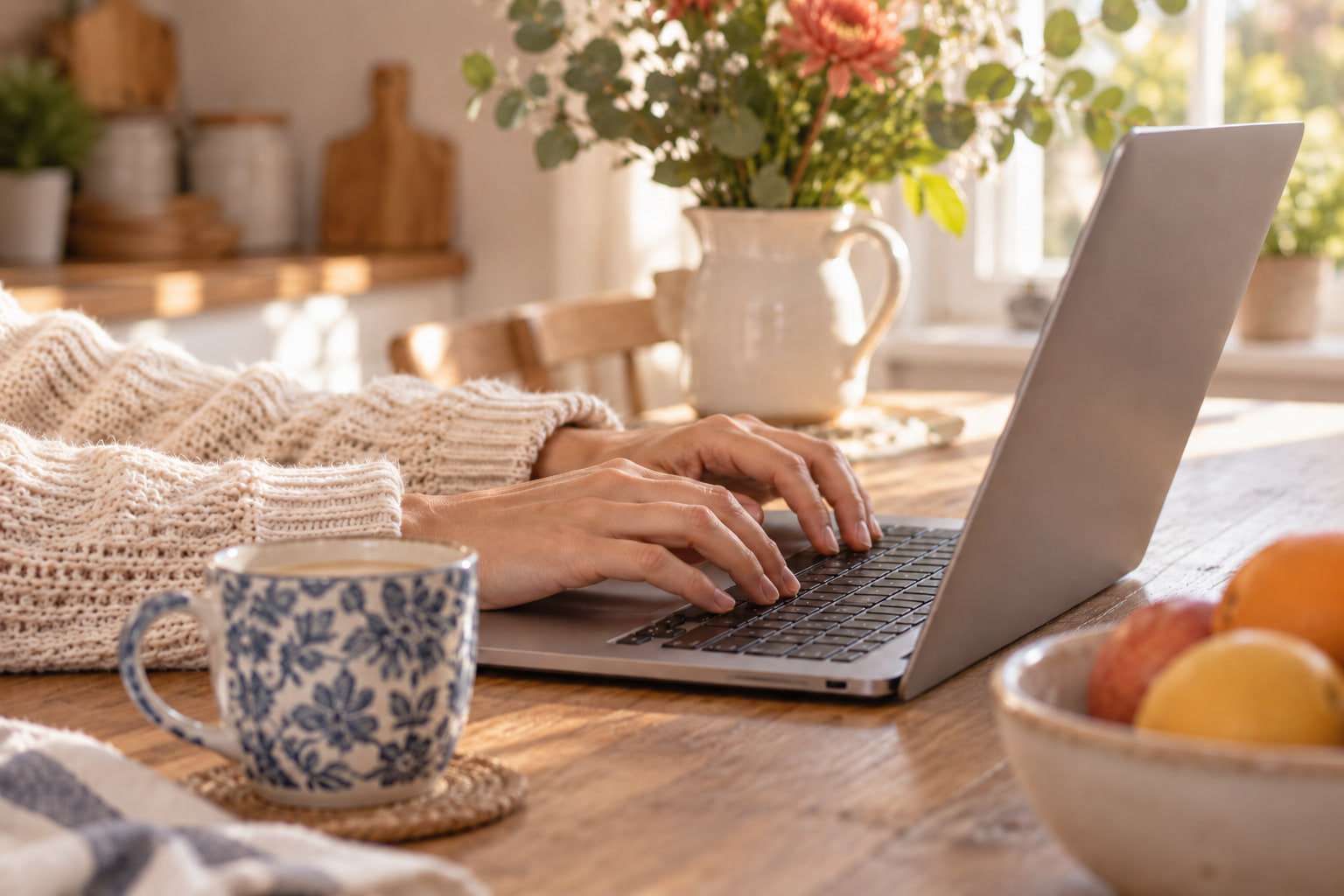 Hands in a cream knitted jumper typing on a laptop at a kitchen table, beside a blue-and-white mug and a bowl of fruit.