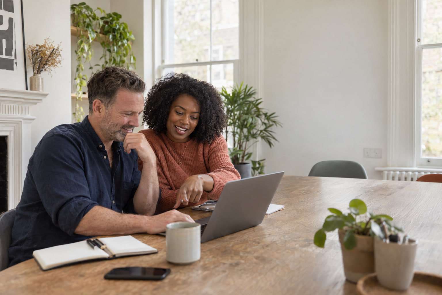 Two colleagues side by side at a long table in a bright office, looking at a laptop together and smiling.