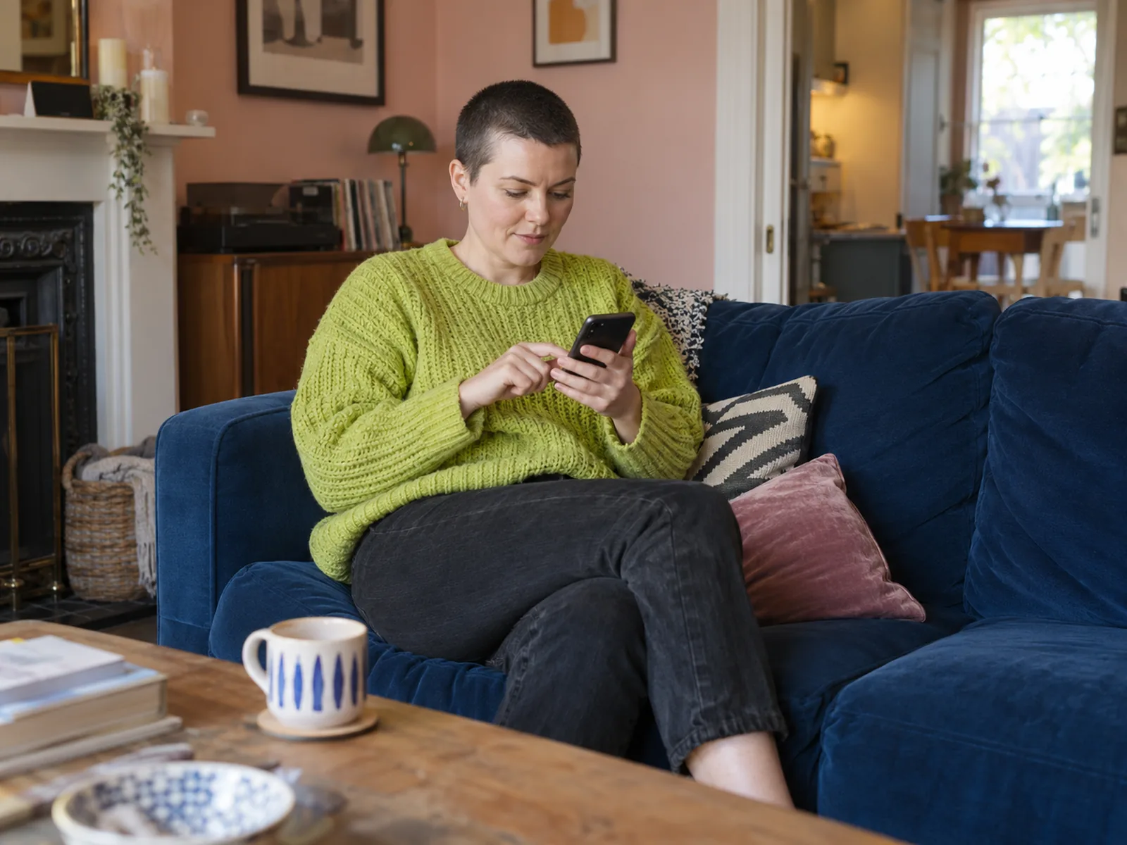 Woman with a buzz cut and a lime jumper using her phone on a blue sofa