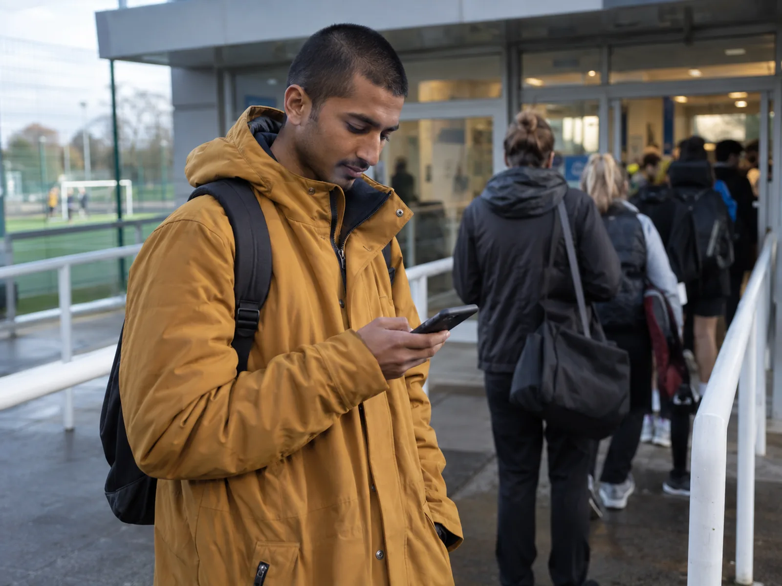 Young man in an amber parka reading his phone in a queue outside a sports centre
