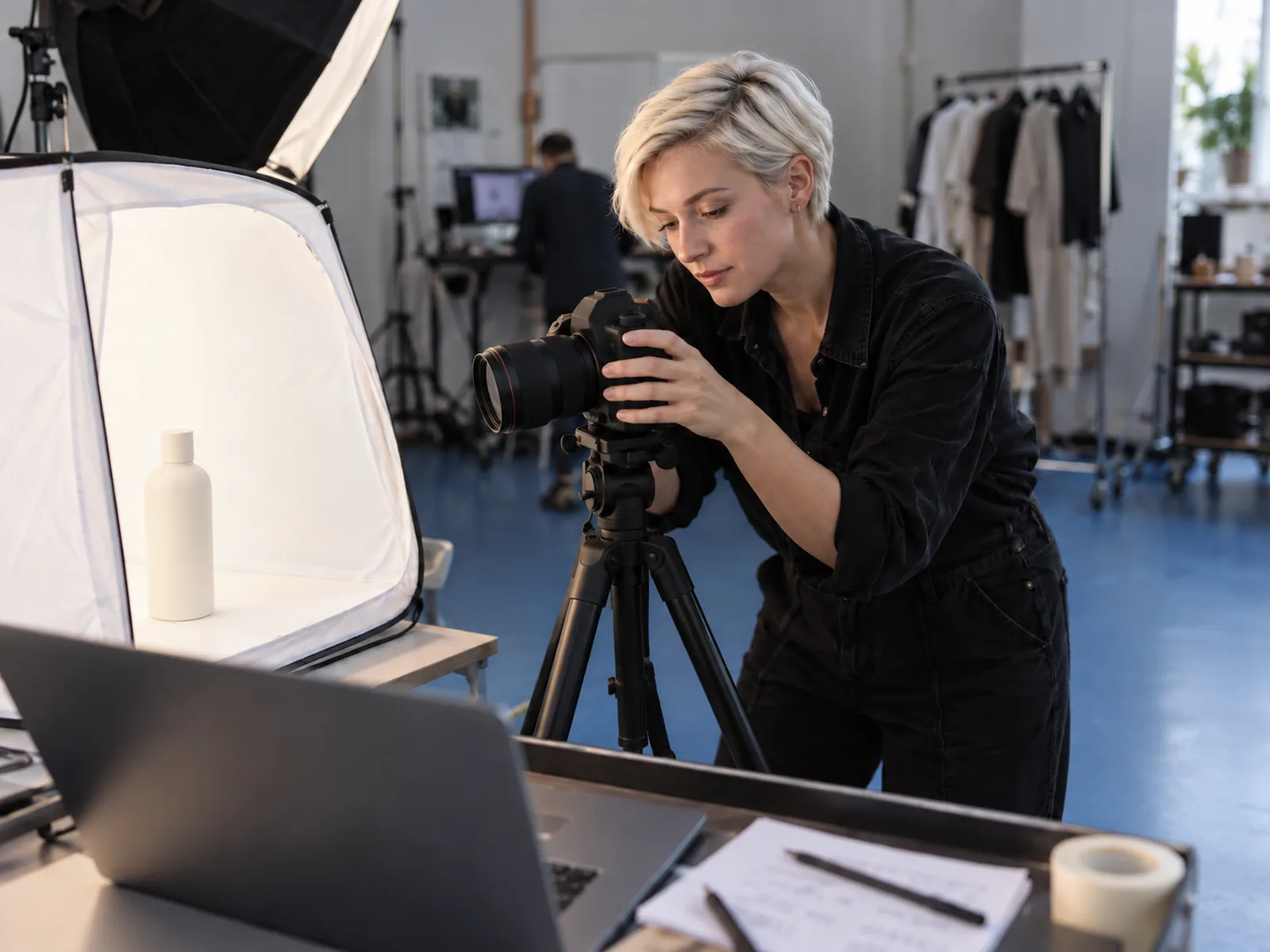 Woman with cropped platinum hair photographing a plain bottle in a white light tent