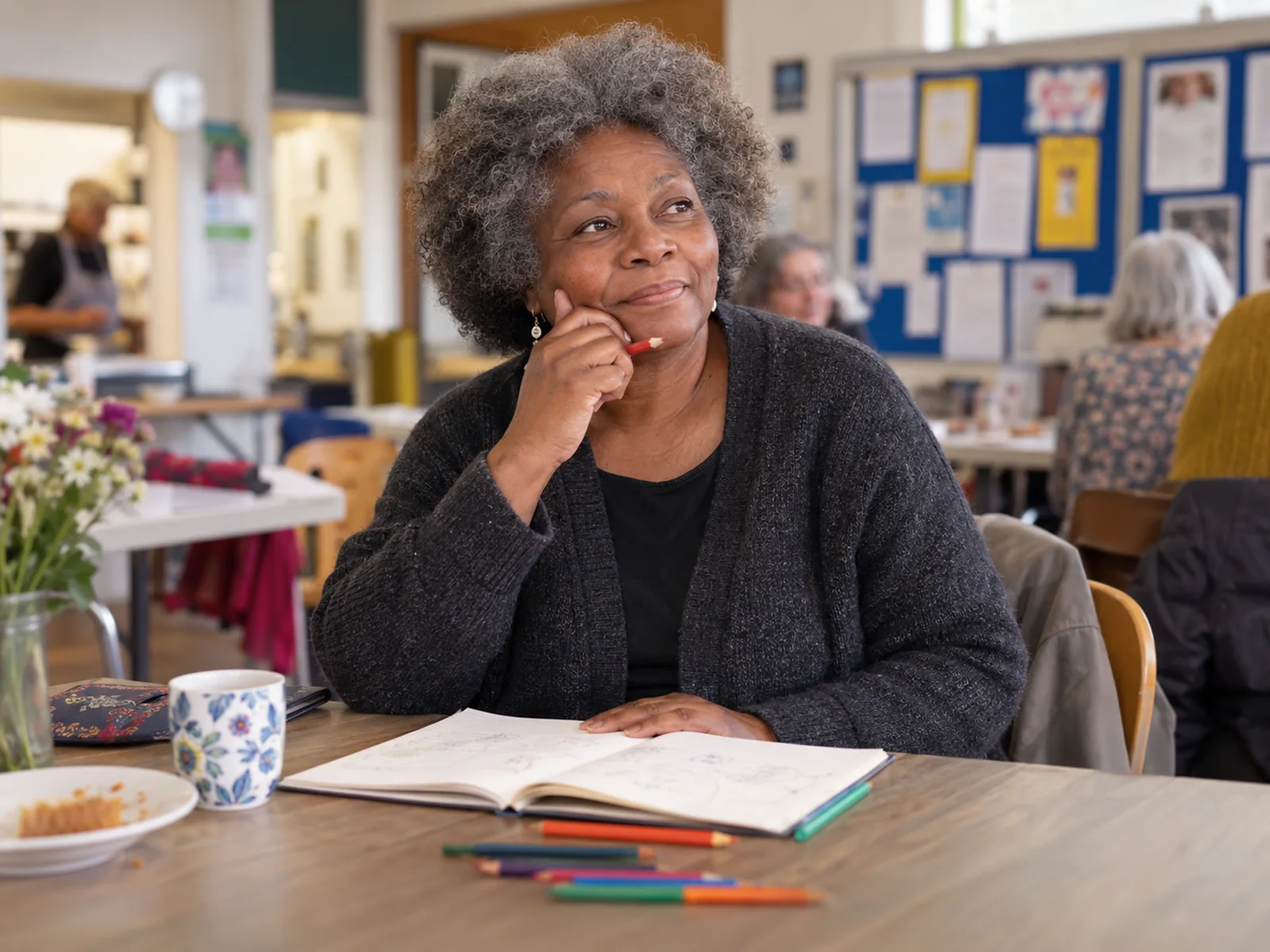 Woman with a salt-and-pepper afro thinking over a notebook at a table in a community hall