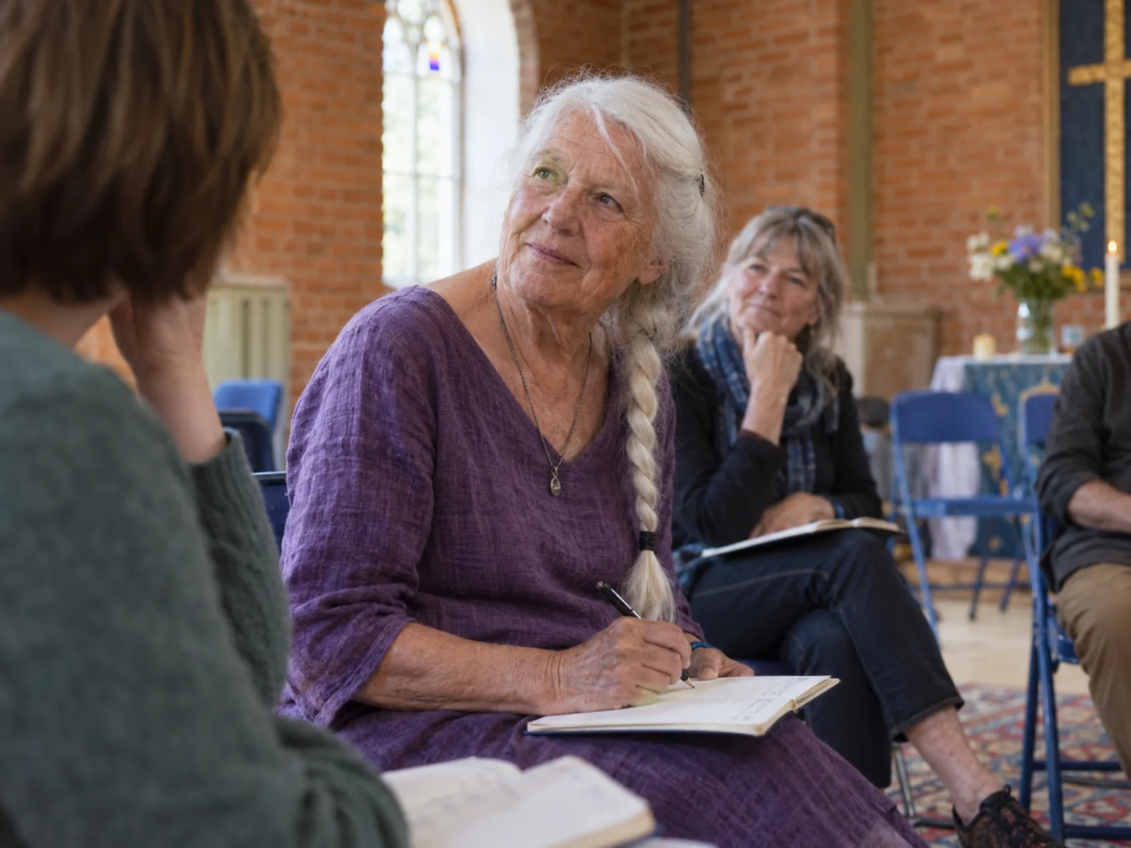 Older woman with a long white plait taking notes during a small group discussion in a brick hall