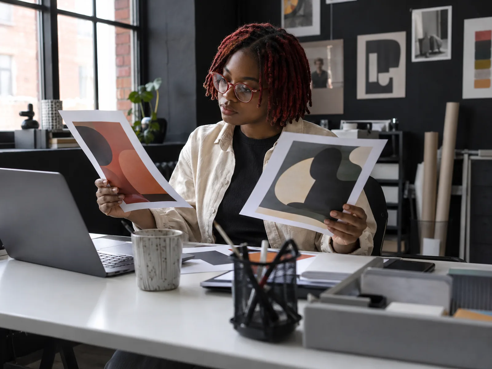 Designer with red locs and round glasses comparing two printed mock-ups in a dark studio