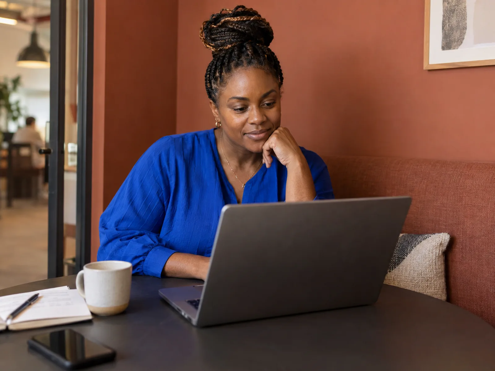 Woman with a braided bun in a cobalt top reading a laptop in a terracotta meeting booth