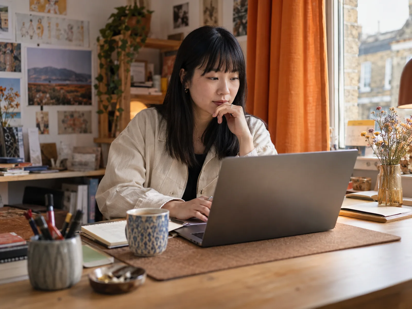 Woman with a black fringe thinking at a laptop in a small studio with orange curtains
