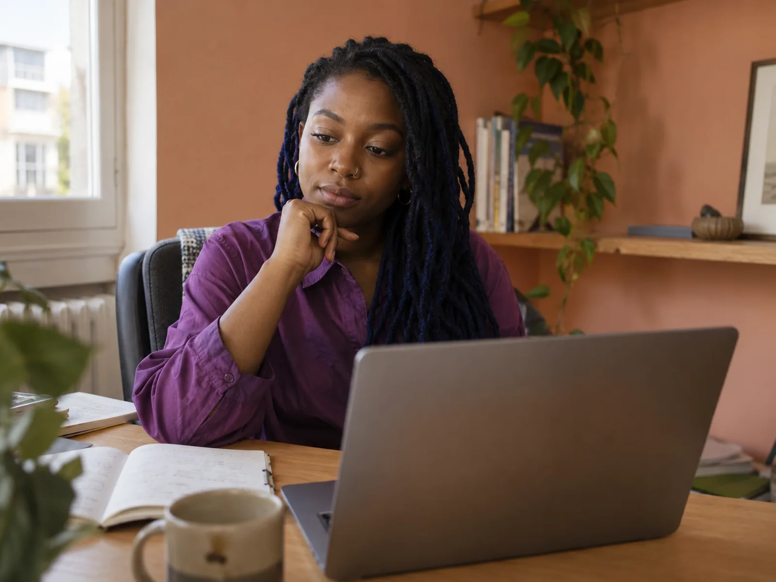 Woman with long locs and a violet shirt pausing over a laptop in a peach-walled alcove