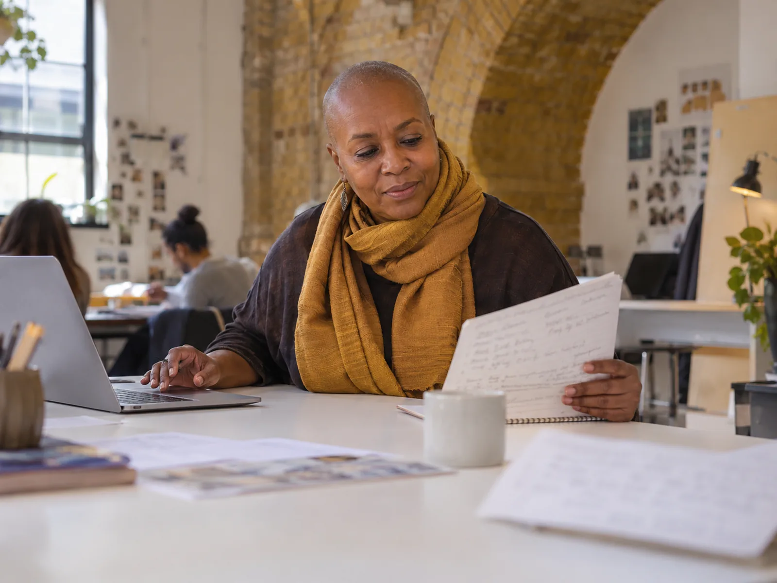 Woman with a shaved head and a golden scarf reading handwritten notes beside a laptop