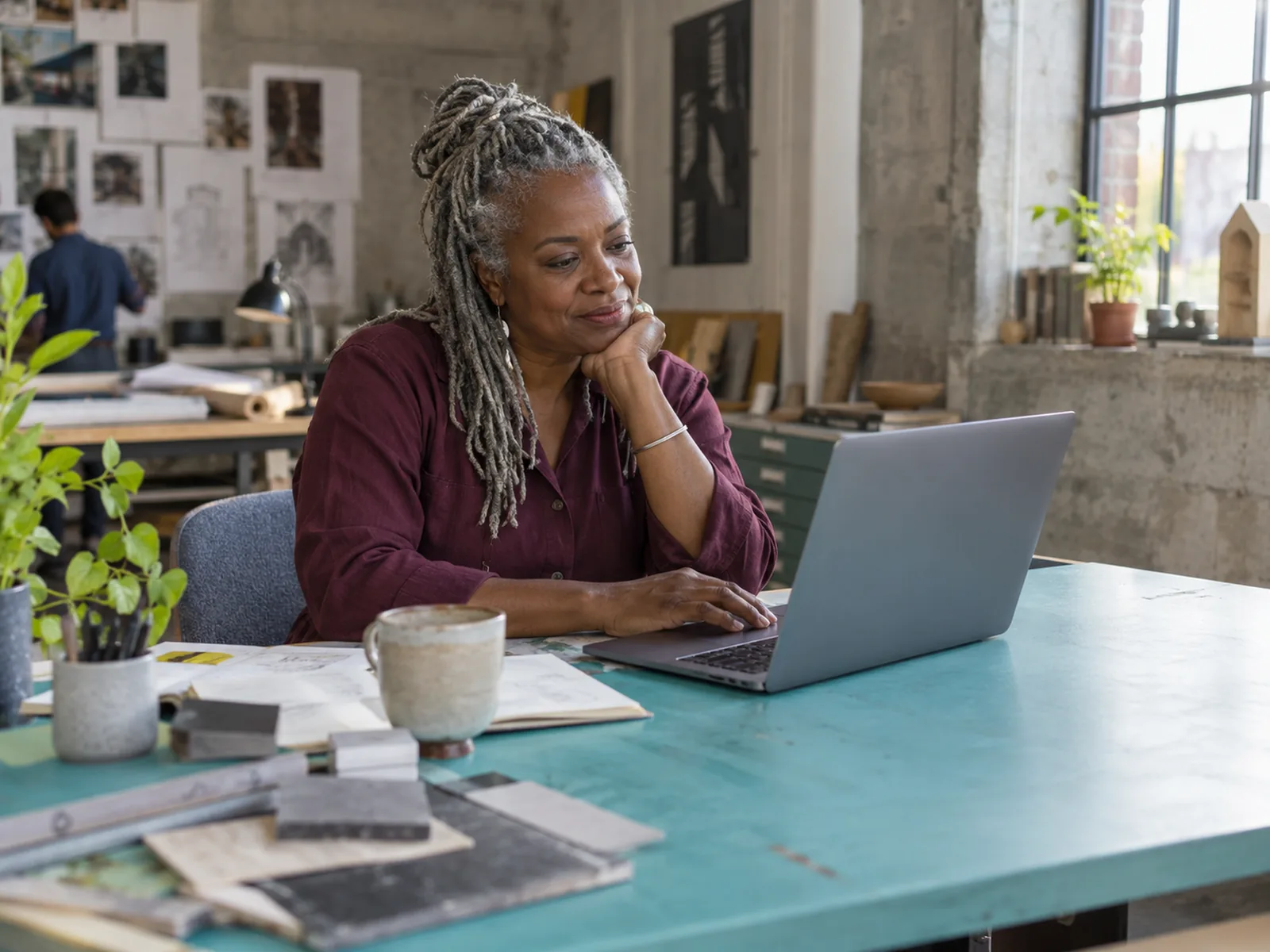 Woman with silver locs looking at a laptop at a turquoise desk in a design studio