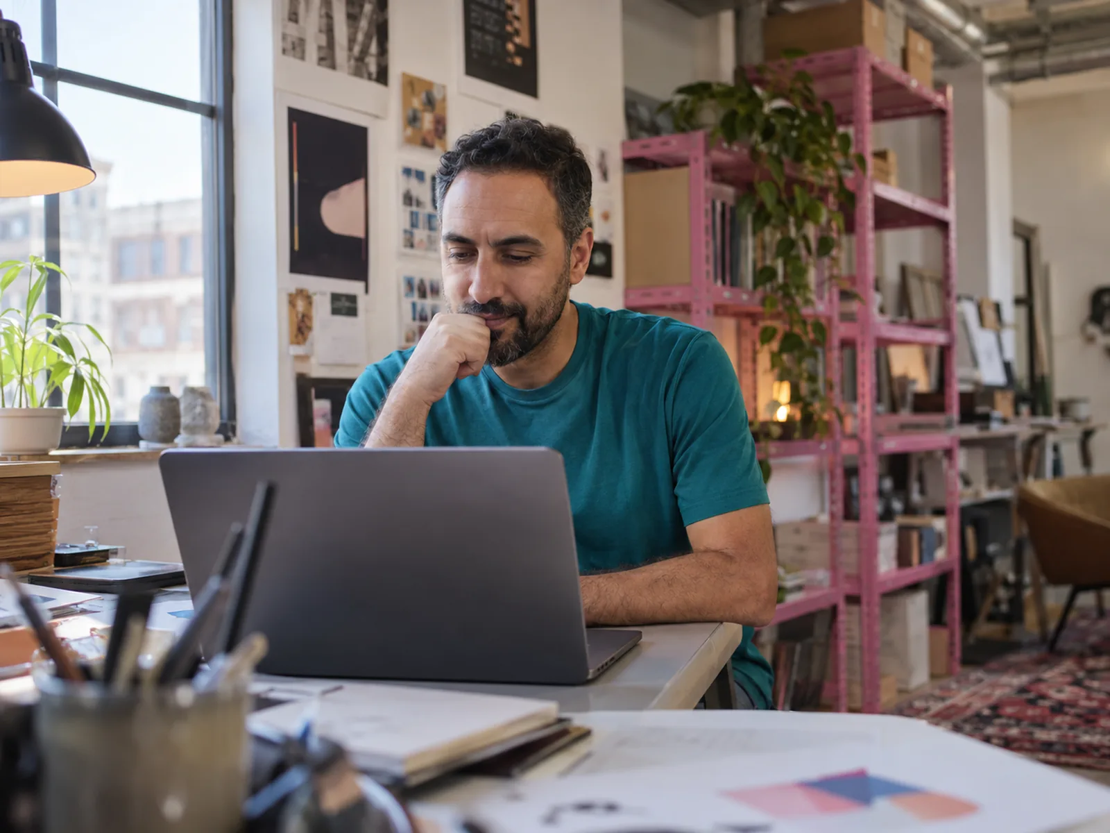 Man with a trimmed beard in a teal T-shirt studying a laptop by pink metal shelving