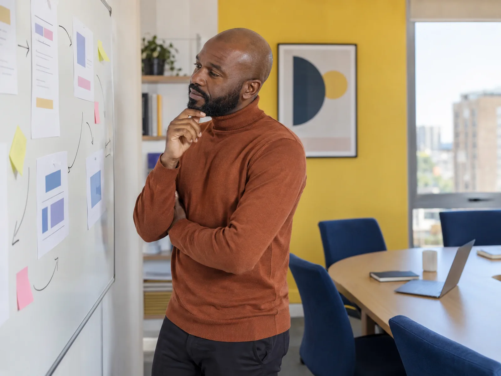 Man in a terracotta jumper thinking at a whiteboard covered in sticky notes