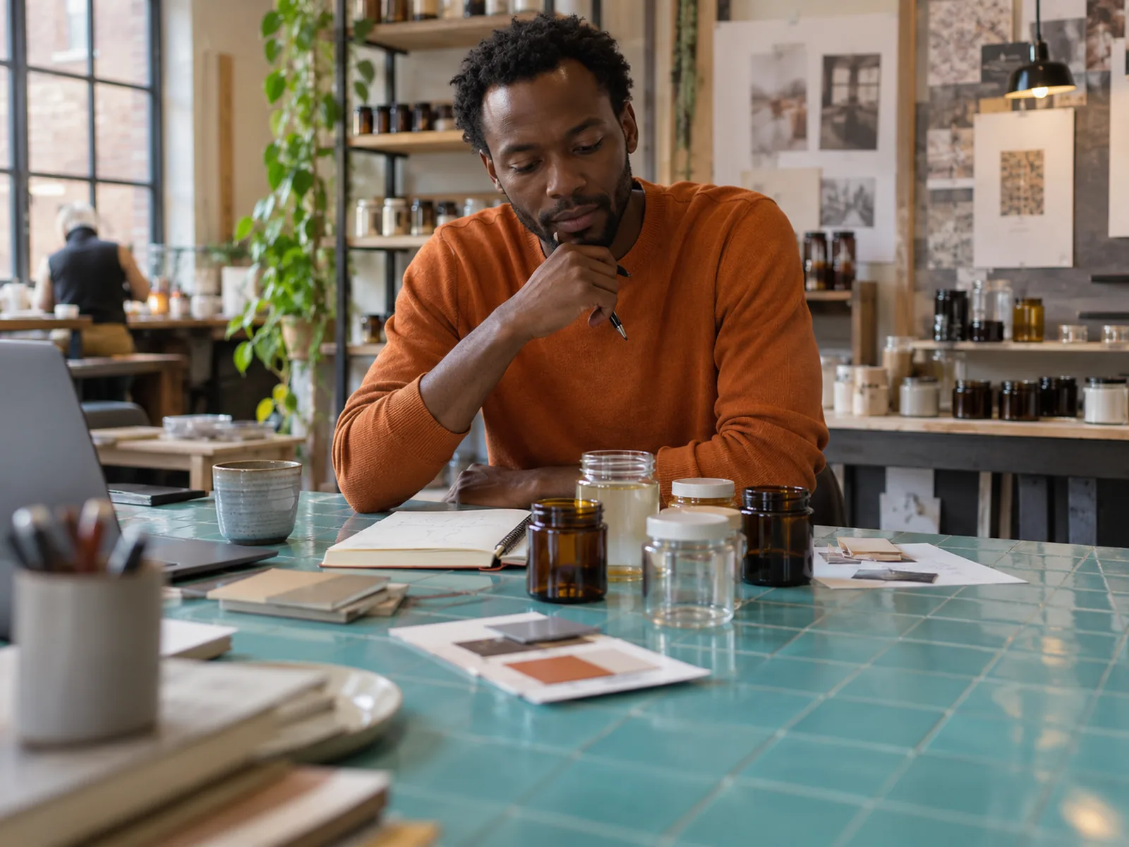 Man in an orange jumper thinking over small jars at a turquoise tiled worktable