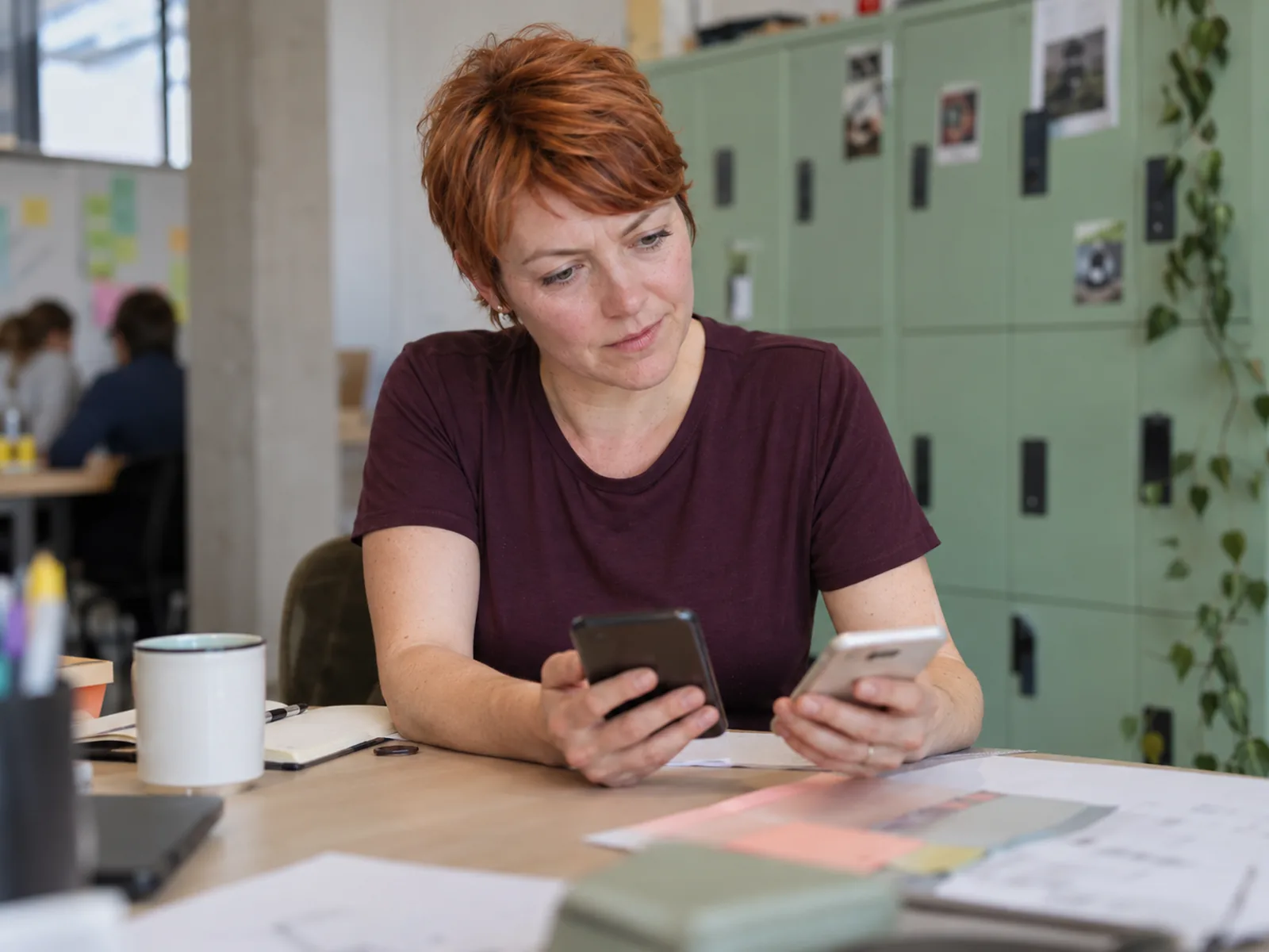 Woman with a ginger pixie cut comparing two phones at a desk near green lockers
