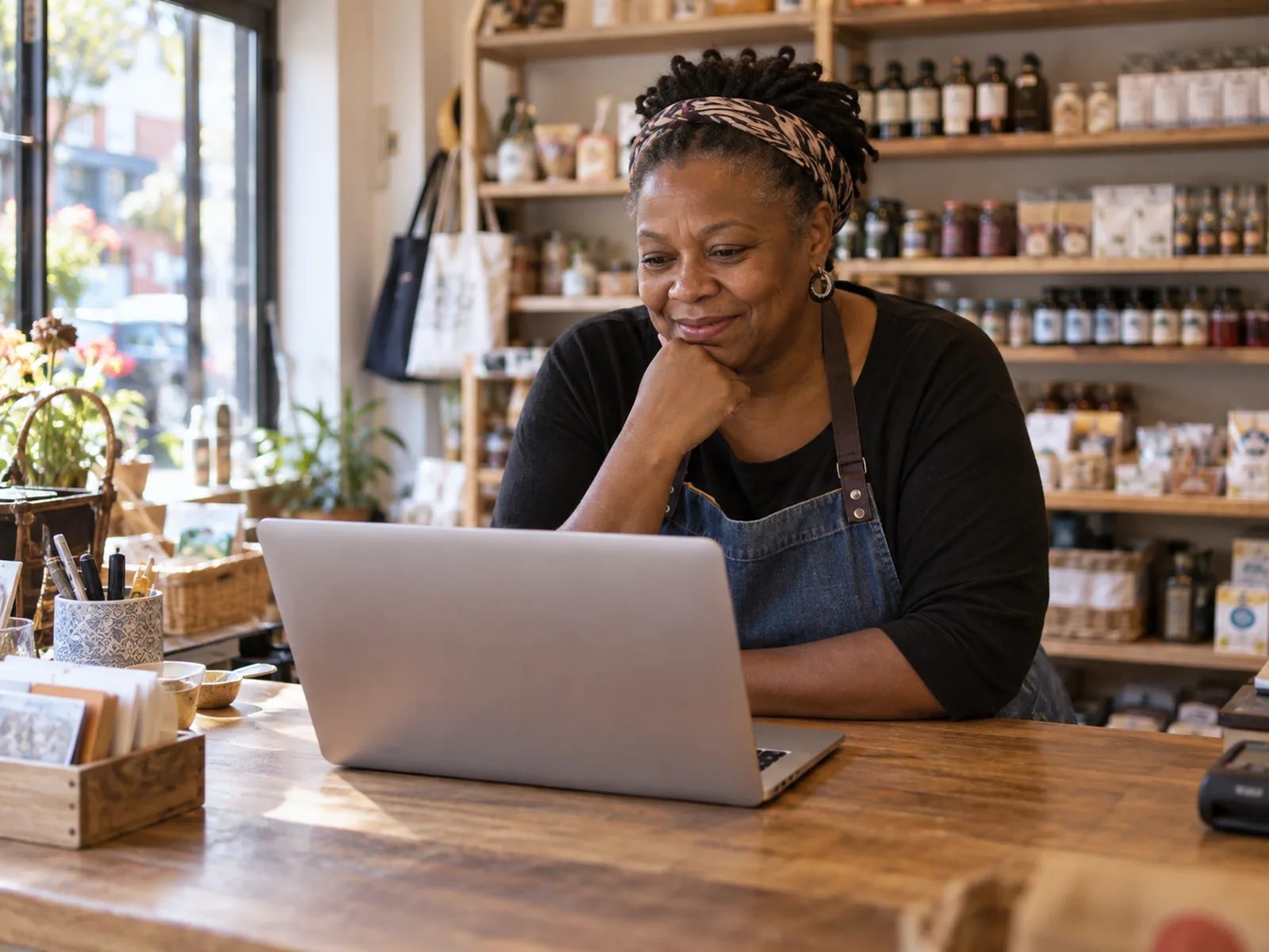Woman with a headscarf smiling at a laptop on the counter of a small food shop
