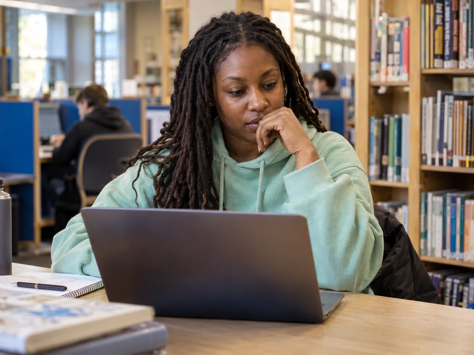 Woman with long locs in a mint hoodie working on a laptop in a library