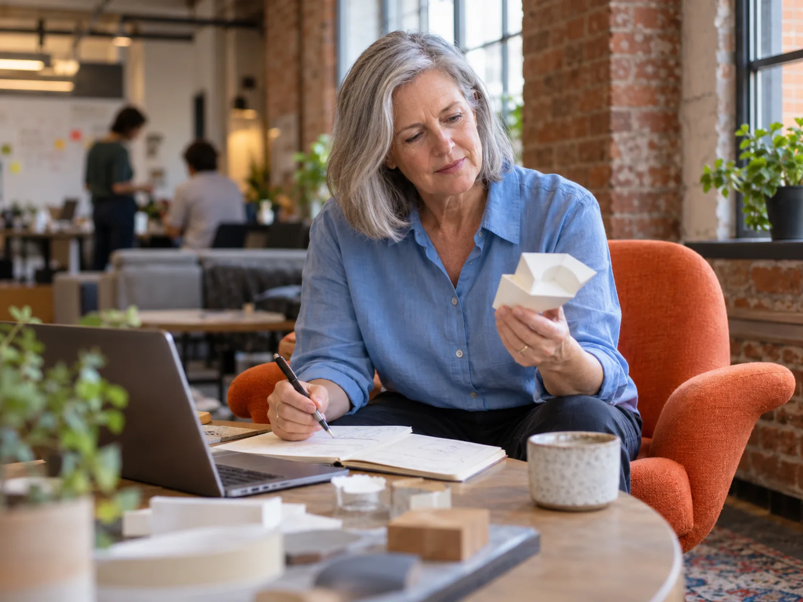 Woman with grey hair examining a small paper prototype in an armchair beside a laptop