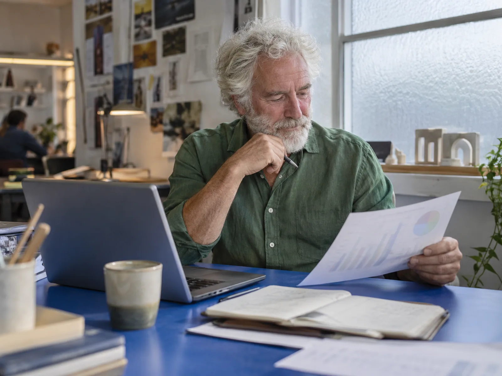Older man with a white beard reading printed charts beside a laptop at a blue desk