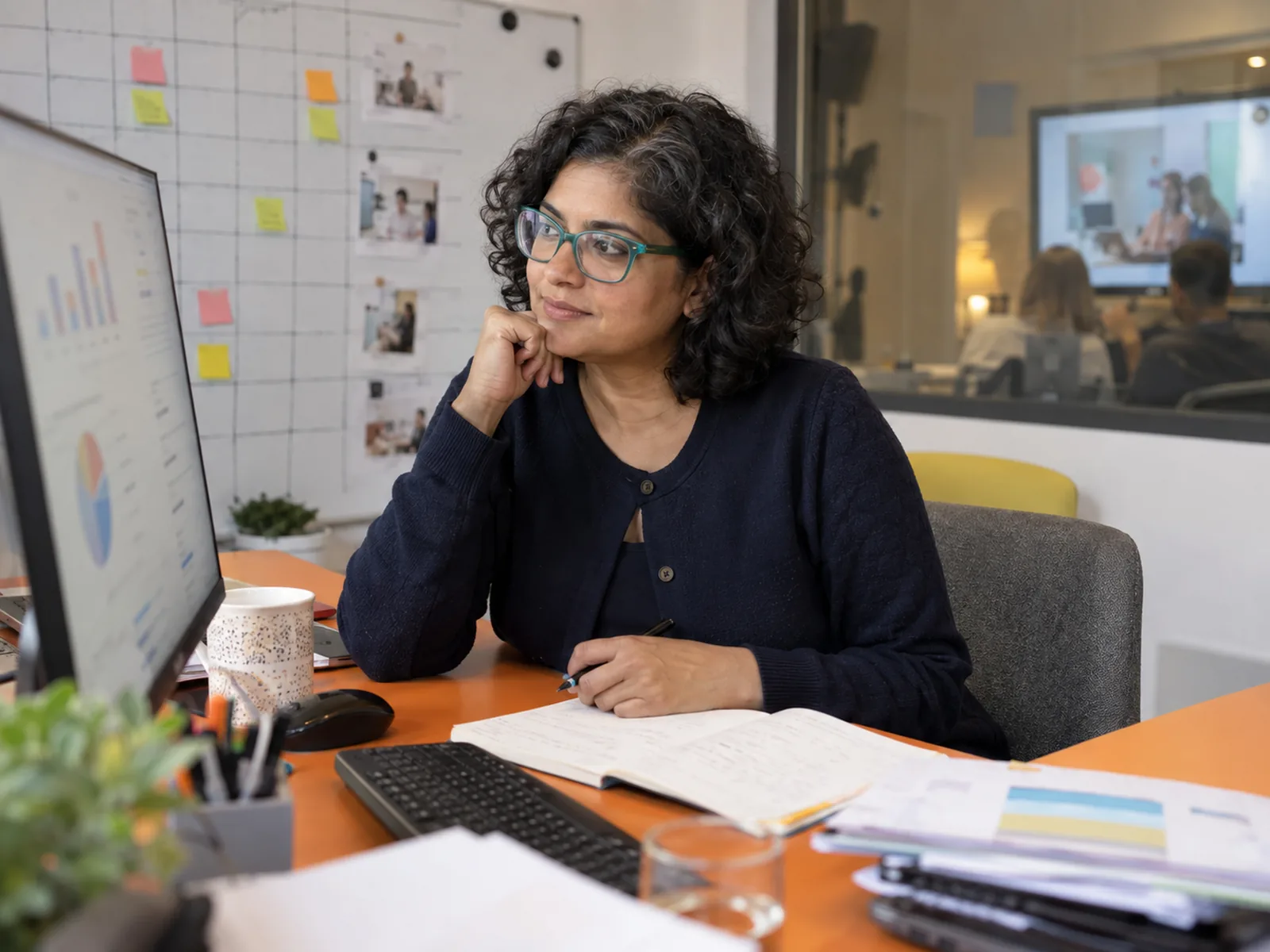 Woman with a curly bob and turquoise glasses studying a monitor at an orange desk