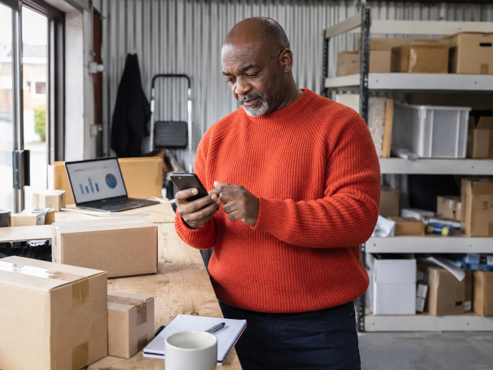 Man with a grey goatee in an orange jumper checking his phone beside parcels in a stockroom