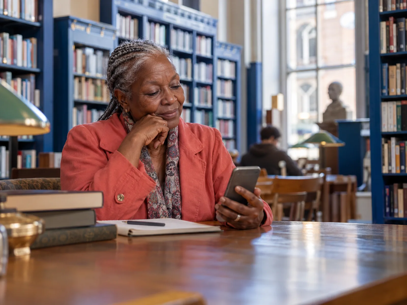 Older woman with silver cornrows and a coral jacket reading her phone in a library