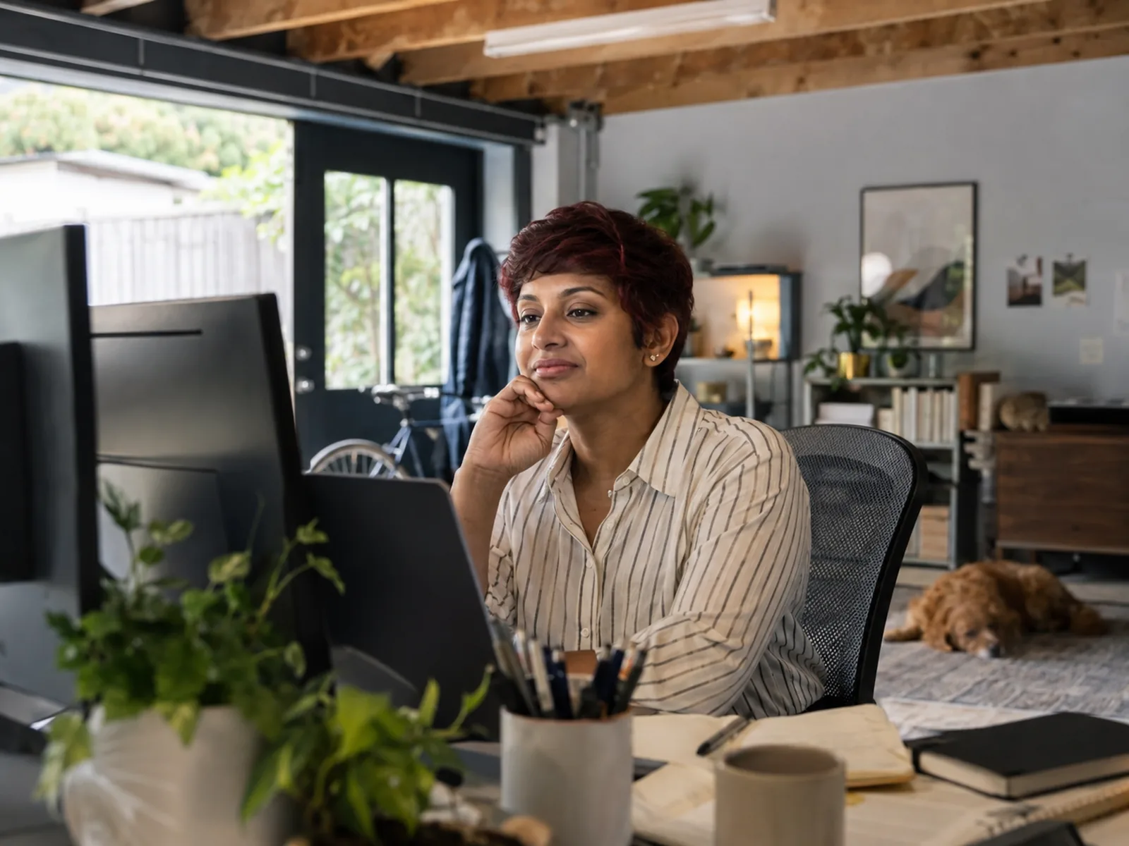 Woman with a dark-red pixie cut working at two monitors in a converted garage office