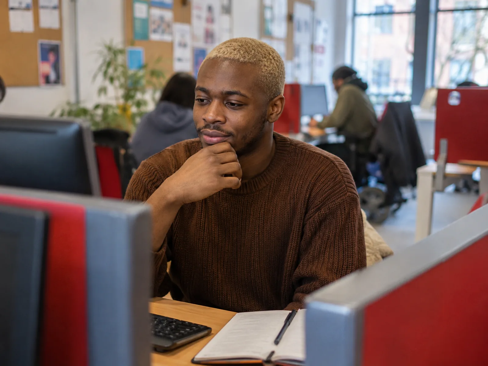 Young man with dyed blond hair thinking at a desktop computer in a community digital hub