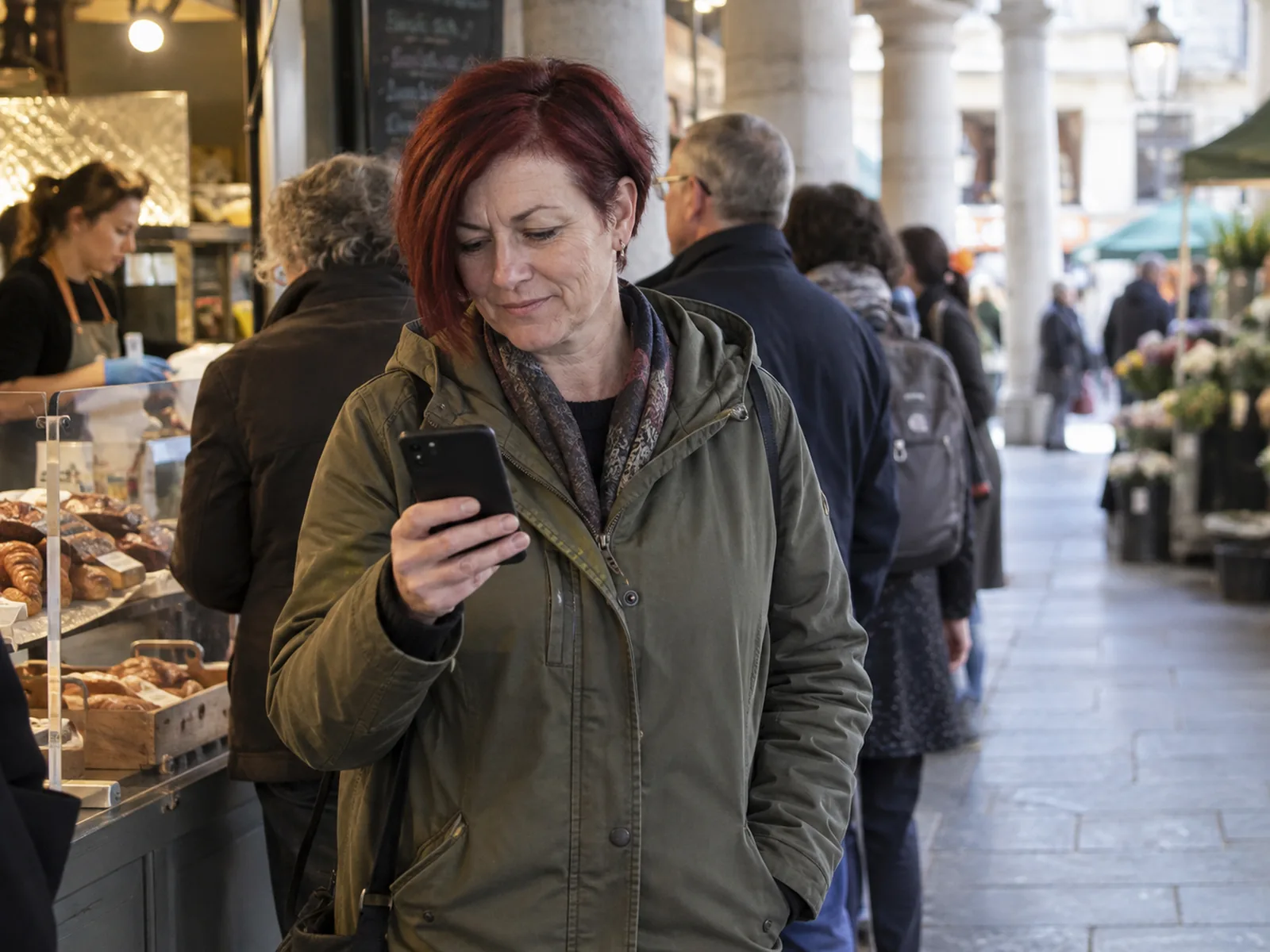 Woman with a dark-red bob and olive anorak reading her phone in a covered market