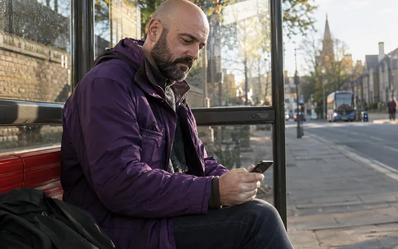 Bald man in a purple jacket checking his phone in a bus shelter