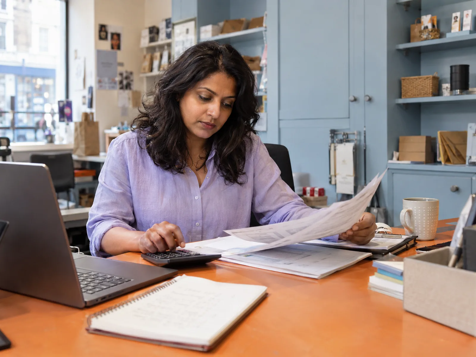 Woman in a lavender shirt reading a printout with a calculator at an orange desk